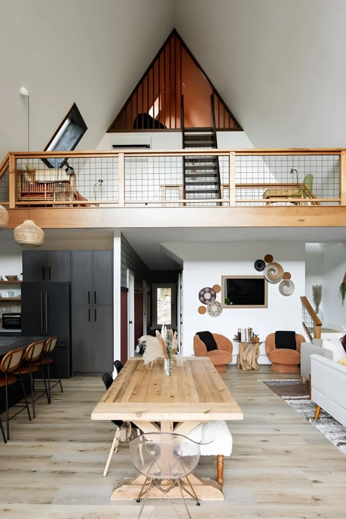 A-frame house interior with an open loft, a rustic wooden dining table, transparent chairs, and natural wood accents creating a warm, modern aesthetic.