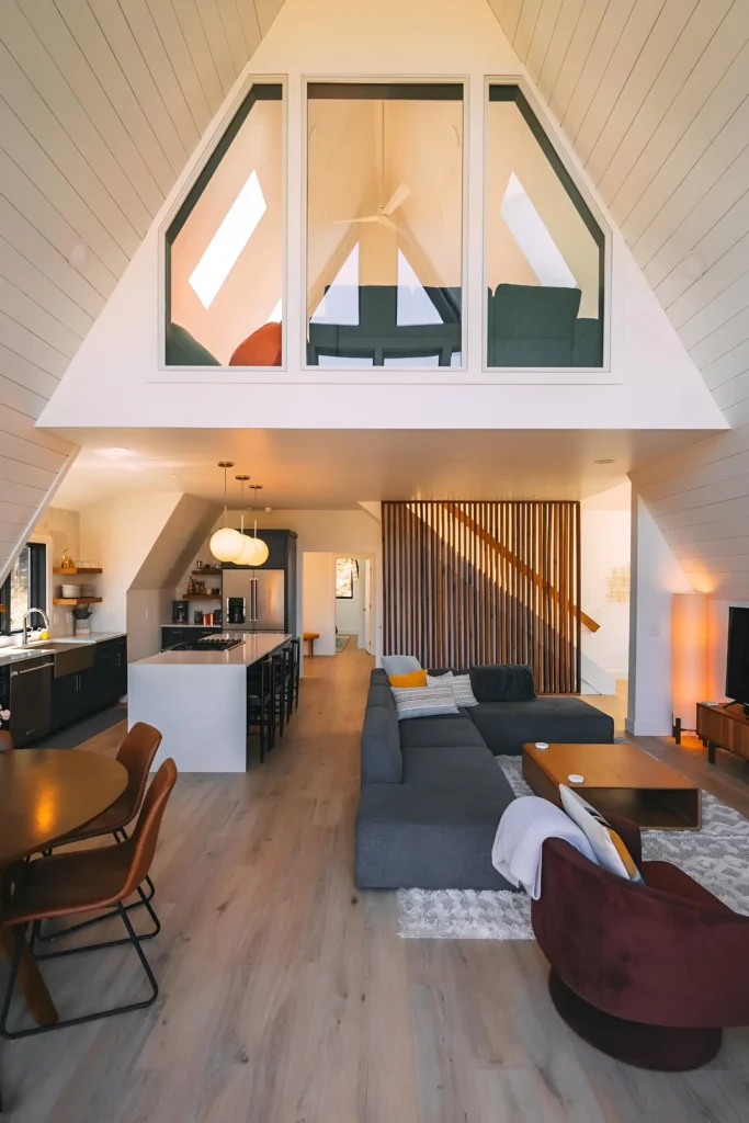 Modern A-frame house interior with a lofted window, dark kitchen cabinetry, a waterfall island, a slatted wood divider, and a cozy living area featuring vibrant accents.