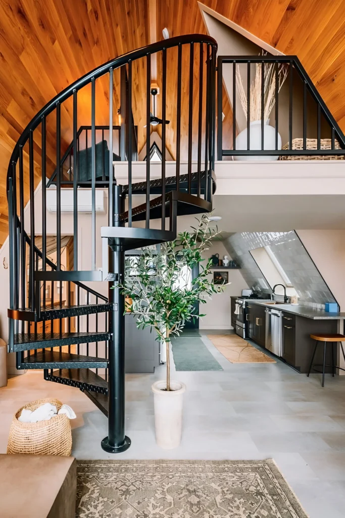 A-frame house interior featuring a black spiral staircase, warm wood-paneled ceiling, modern kitchen with dark cabinetry, and natural accents like a potted plant and woven basket.