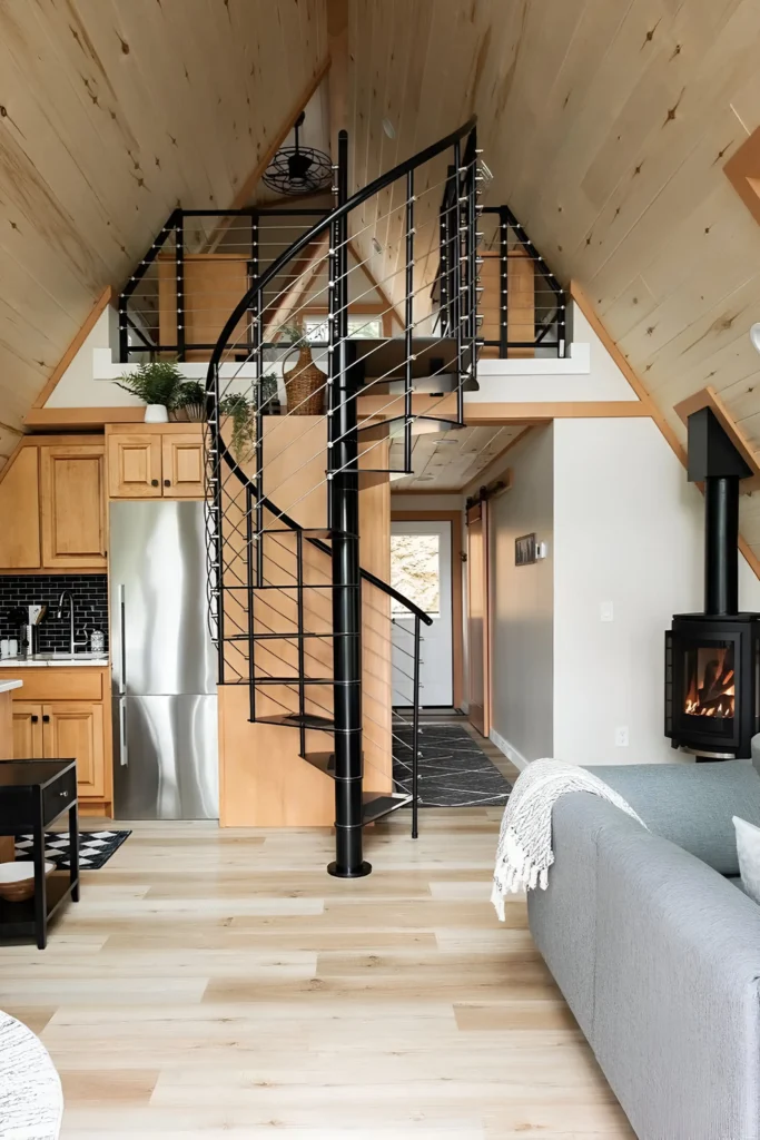 A-frame house interior featuring a blond wood finish, black spiral staircase, modern loft railings, compact kitchen with black subway tile backsplash, and a cozy corner fireplace.