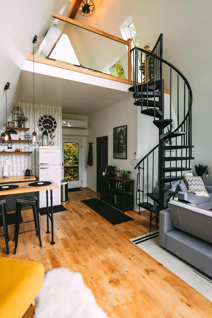 A-frame house interior with a spiral staircase, loft with glass panels, patterned accent wall, vintage-style refrigerator, and light wood flooring accented by a yellow armchair.