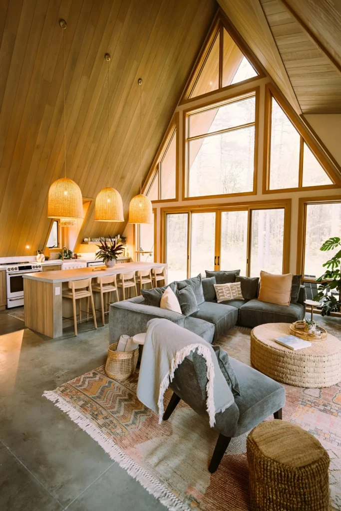 A-frame house interior with natural wood paneling, large triangular windows, a cozy sectional sofa, rattan pendant lights, and an open-concept kitchen with a wooden island.