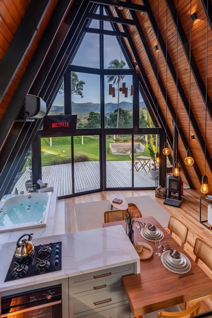 A-frame house interior with floor-to-ceiling windows, a wood-burning stove, pendant lighting, built-in spa tub, modern kitchen with marble countertop, and a dining table overlooking stunning outdoor views.