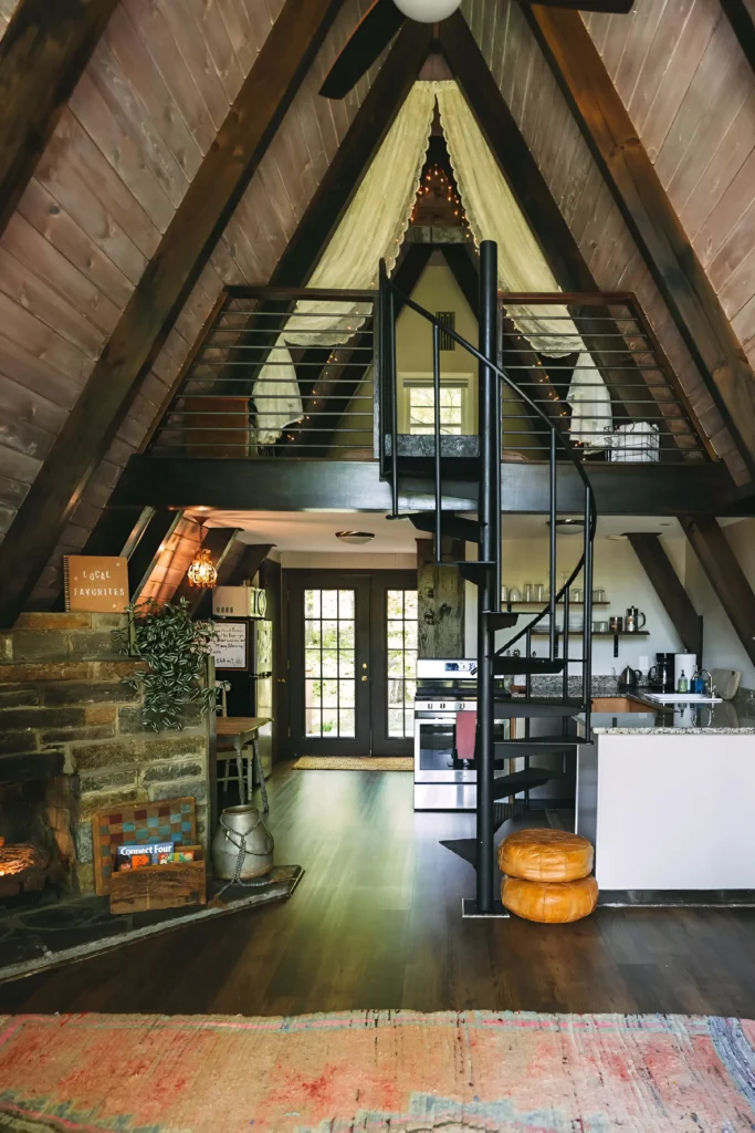 A-frame house interior with dark wood paneling, a stone fireplace, spiral staircase, loft with draped curtains and string lights, compact kitchen with open shelving, and French doors.