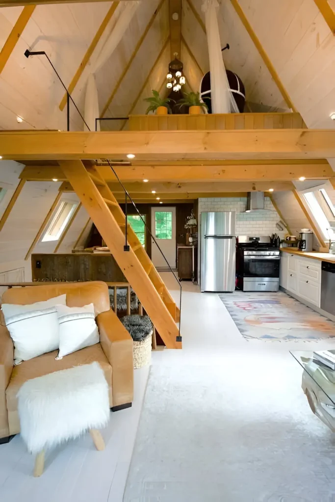 A-frame house interior featuring natural wood beams, a loft with drapes and plants, whitewashed walls, a leather armchair, and a modern kitchen with subway tiles and stainless steel appliances.