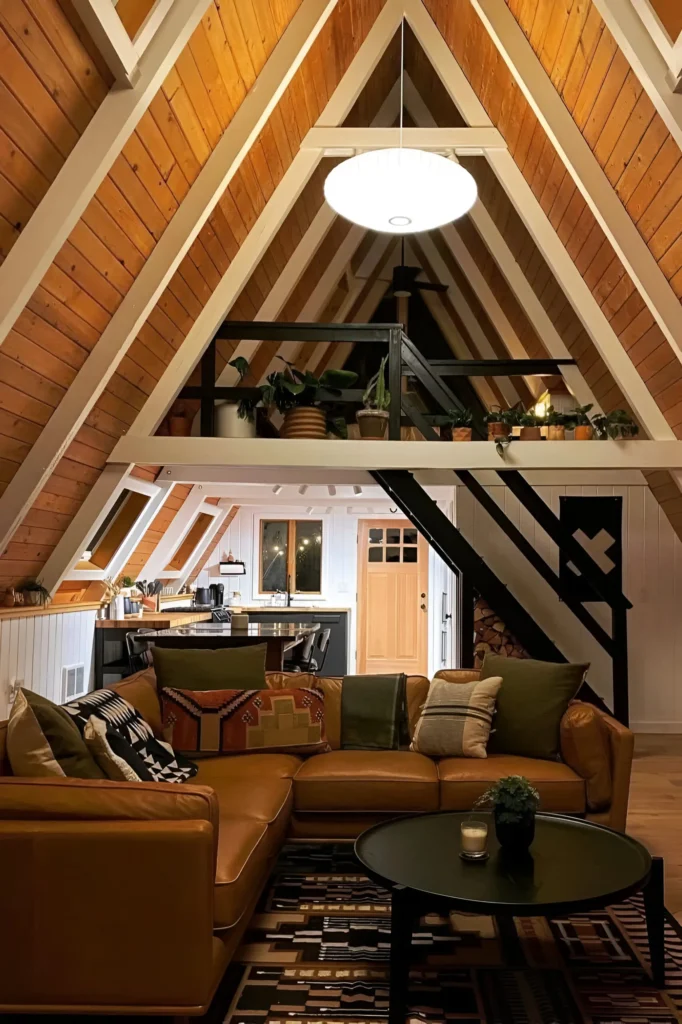 A-frame house interior with rich wood paneling, a tan leather sectional, tribal-inspired textiles, a loft with greenery, and an open kitchen below.