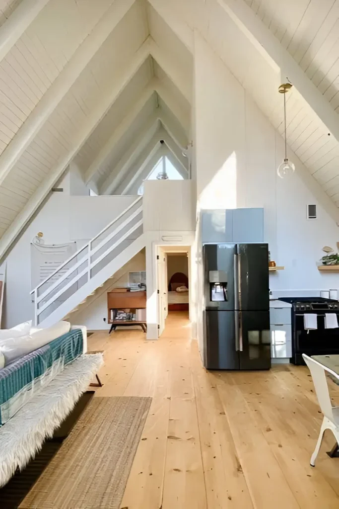 Bright A-frame house interior with white walls, vaulted ceilings, light wood flooring, a modern kitchen, and a staircase leading to a loft.