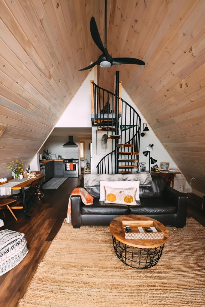 A-frame house interior featuring a black spiral staircase, wooden ceilings and floors, a dark leather sofa, natural woven accents, and a compact kitchen with dark cabinetry.