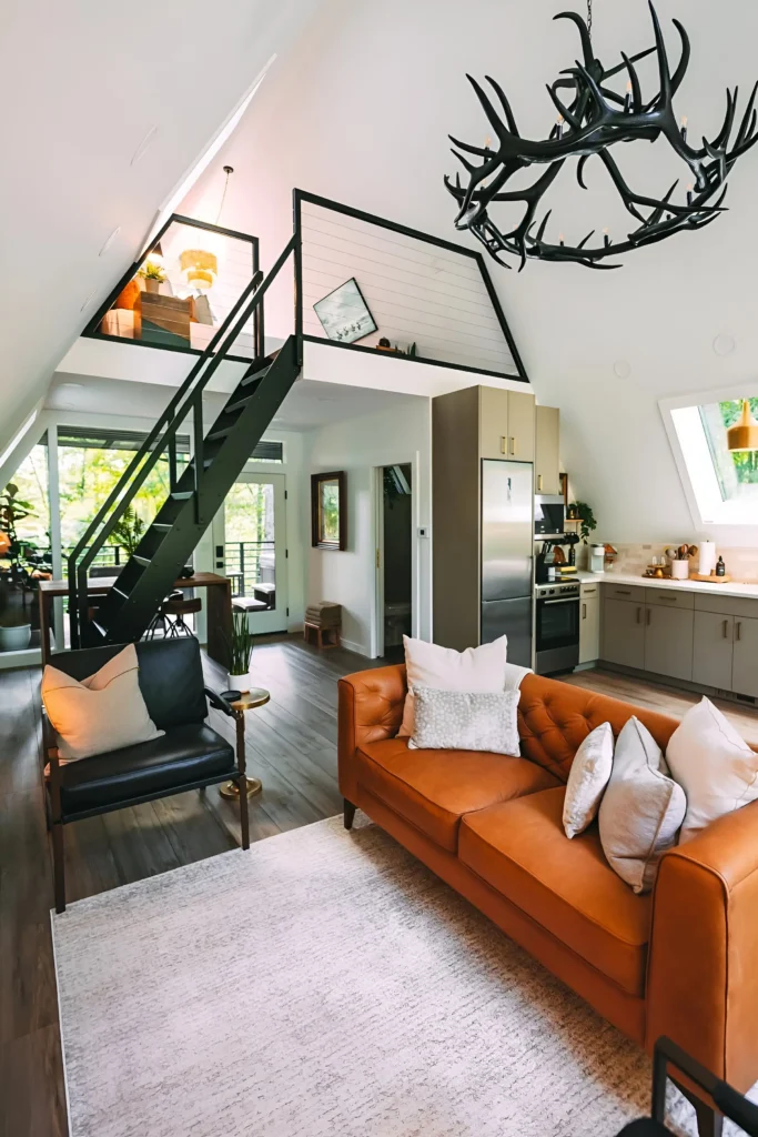 Modern A-frame house interior with a black metal staircase, antler chandelier, warm leather sofa, stainless steel kitchen appliances, and angled windows.