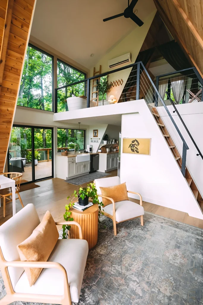 A-frame house interior with floor-to-ceiling windows, a modern kitchen, farmhouse sink, minimalist staircase, light-toned furniture, and loft area with privacy curtains.