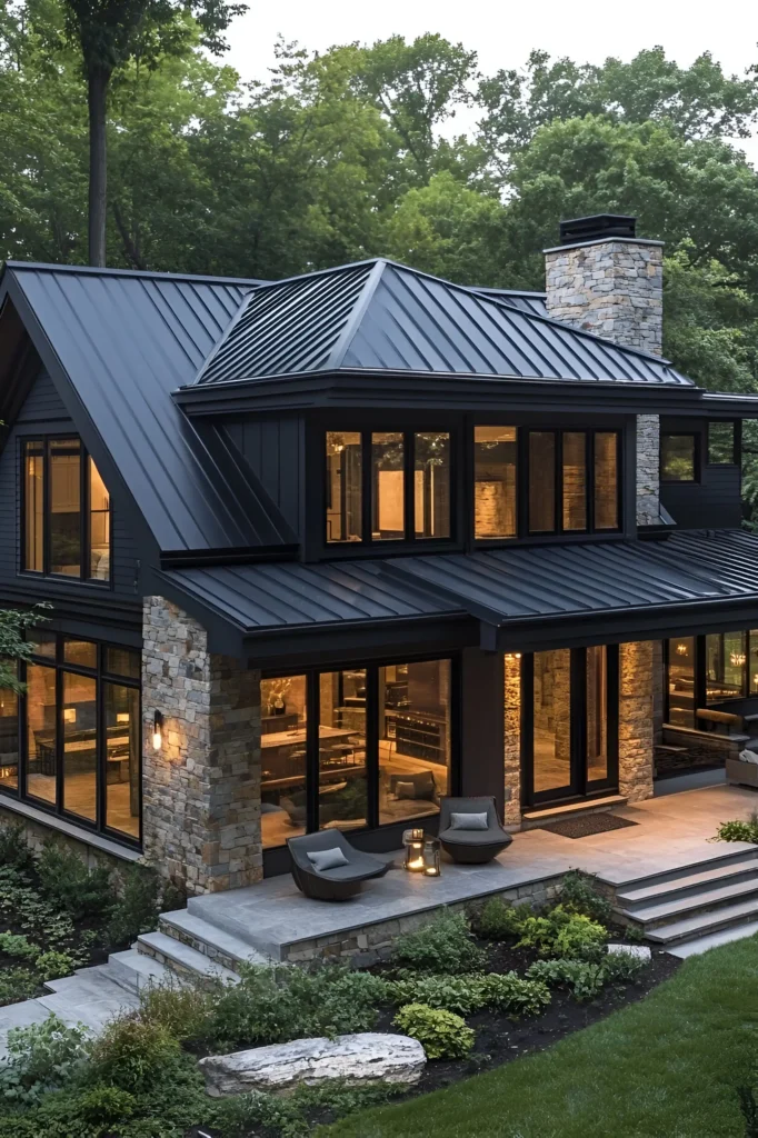 Black farmhouse with a metal roof, stone walls, and large glass windows surrounded by lush greenery, featuring warm interior lighting and outdoor seating on a stone patio.