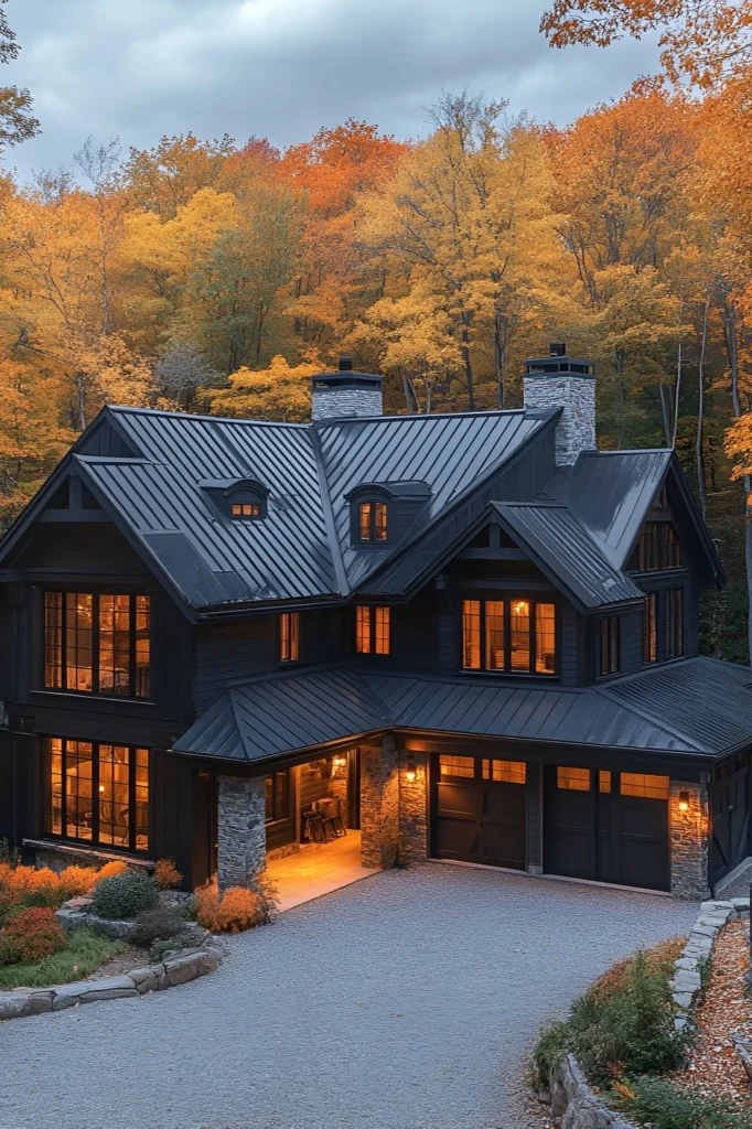 Black farmhouse with a sleek metal roof, stone accents, and warm glowing windows, set against vibrant autumn trees and a gravel driveway.