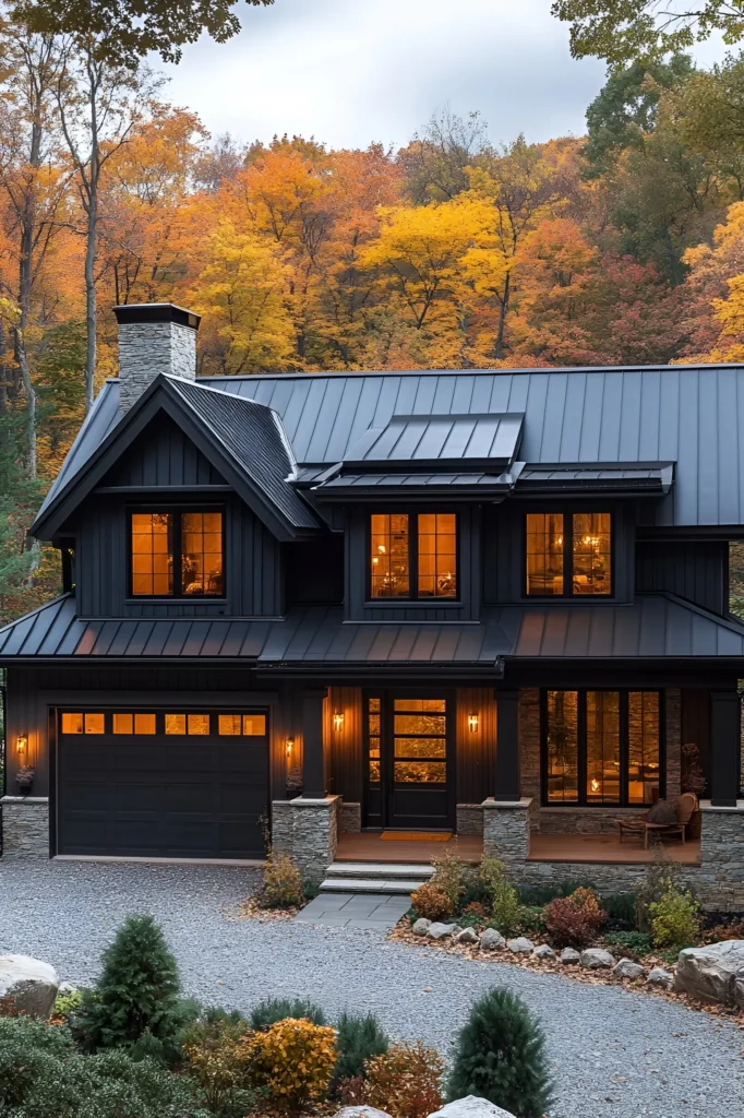 Black farmhouse with a metal roof, stone accents, and glowing windows, surrounded by autumn trees, a gravel driveway, and a landscaped yard.