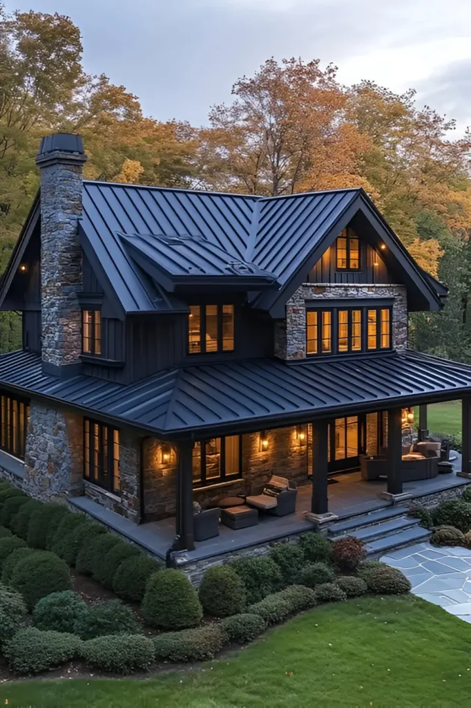 Black farmhouse with a metal roof, stone walls, and a wraparound porch, illuminated by warm lighting, surrounded by manicured greenery in a tranquil countryside setting.