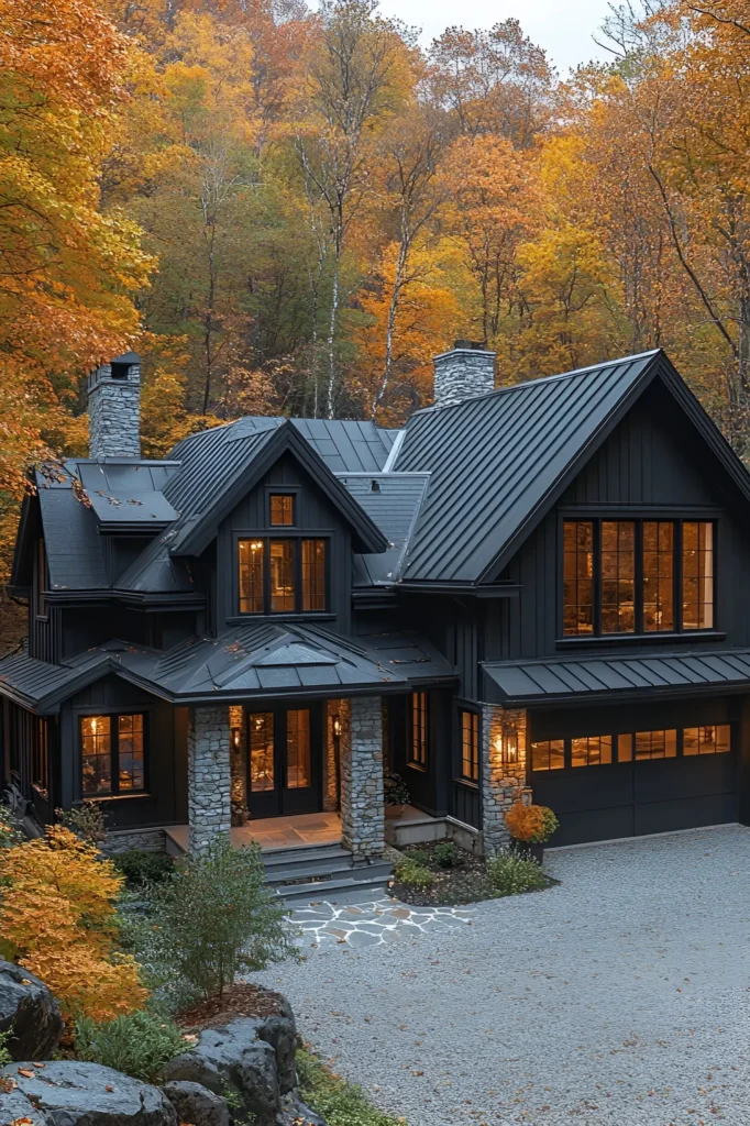 Black farmhouse with stone pillars, a metal roof, and warm-lit windows, set amidst colorful autumn trees with a gravel driveway in a tranquil rural landscape.