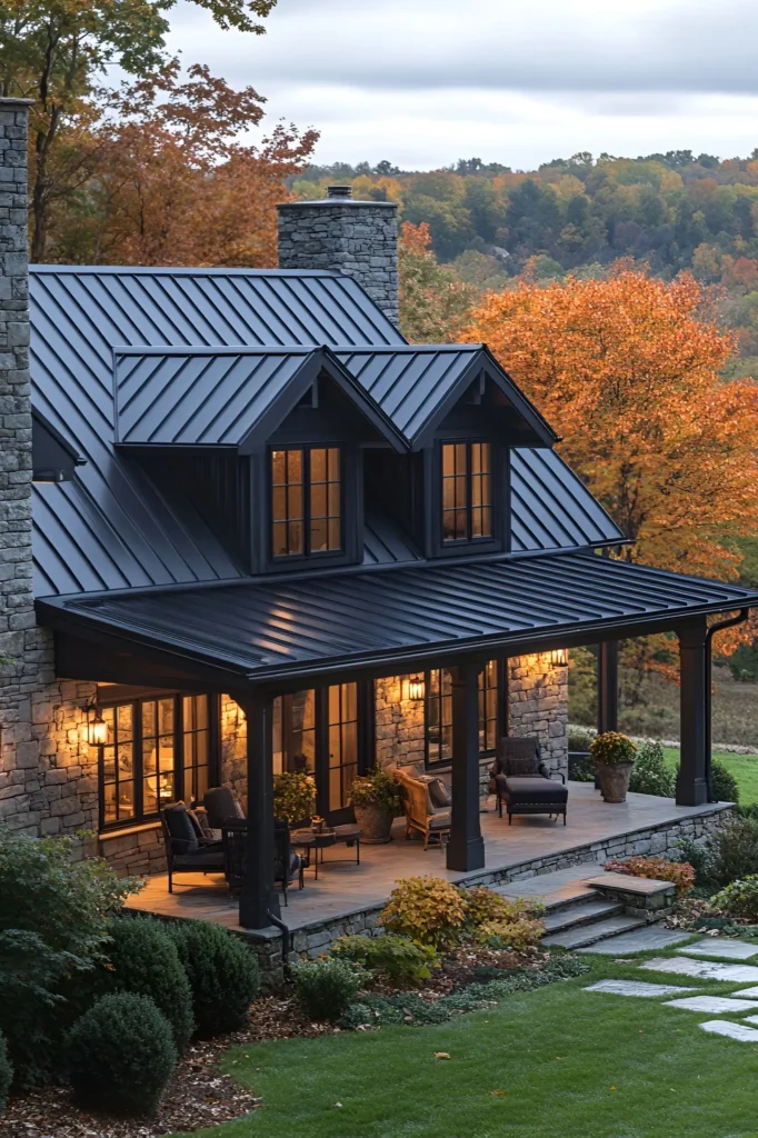 Black farmhouse with a metal roof, stone facade, and a cozy wraparound porch featuring warm lighting and seating, set against autumn foliage and landscaped greenery.