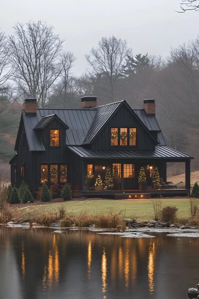 Black farmhouse with a metal roof and festive holiday lights reflecting on a nearby lake, surrounded by misty trees and seasonal greenery in a peaceful countryside setting.