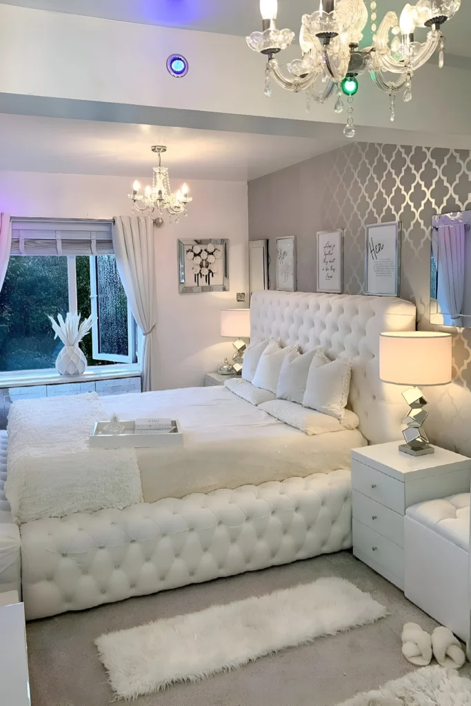 White bedroom with tufted headboard, chandeliers, mirrored decor, and cozy ambiance.