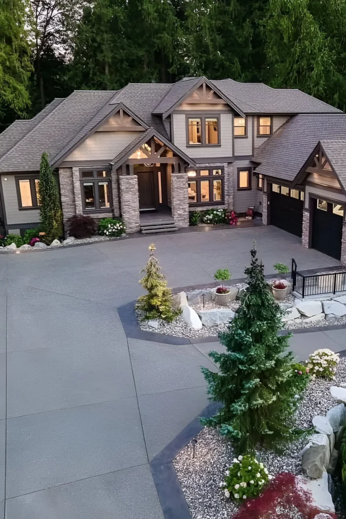 Modern luxury home with stone pillars, gabled roofs, warm lighting, and a landscaped driveway surrounded by trees.