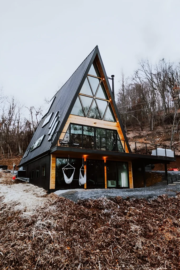 Modern A-frame house with black metal roof, geometric windows, natural wood accents, balcony, and hanging chairs, located on a hillside.