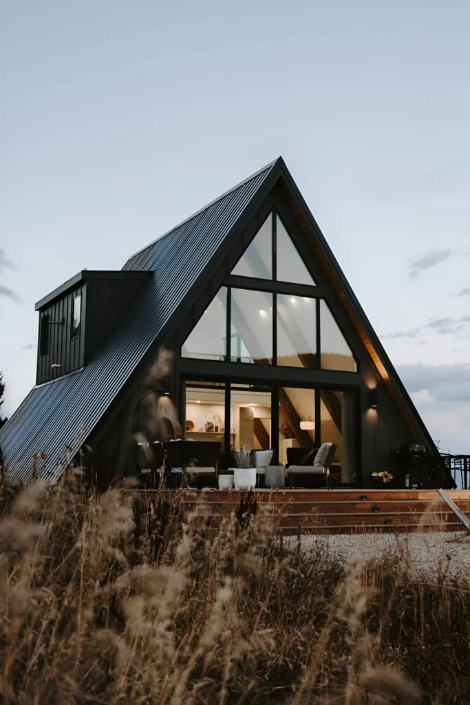 Minimalist A-frame house with corrugated metal roof, large glass windows, warm interiors, and a spacious outdoor deck surrounded by natural grasses.