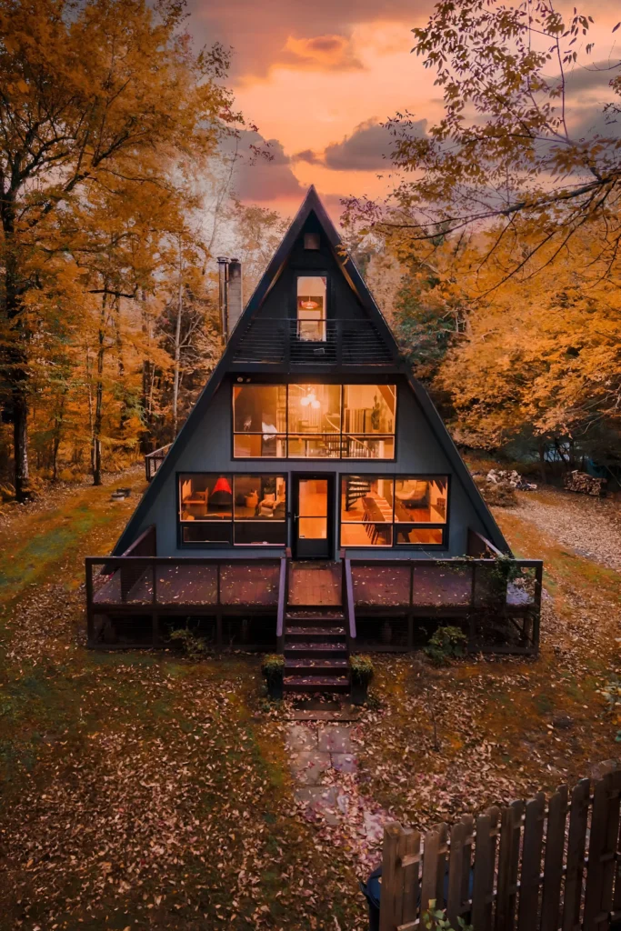 Modern A-frame house with large windows and warm interior lighting, surrounded by golden autumn trees and an expansive deck.