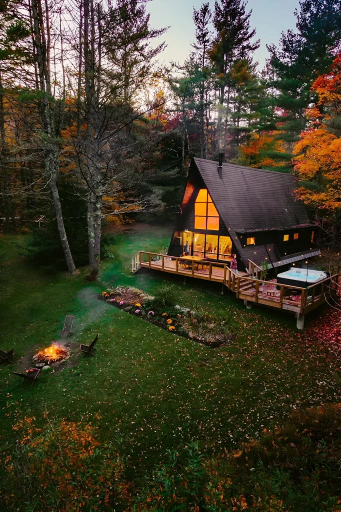 Modern A-frame house with an illuminated deck, hot tub, fire pit, and lush garden, surrounded by a vibrant forest during autumn.