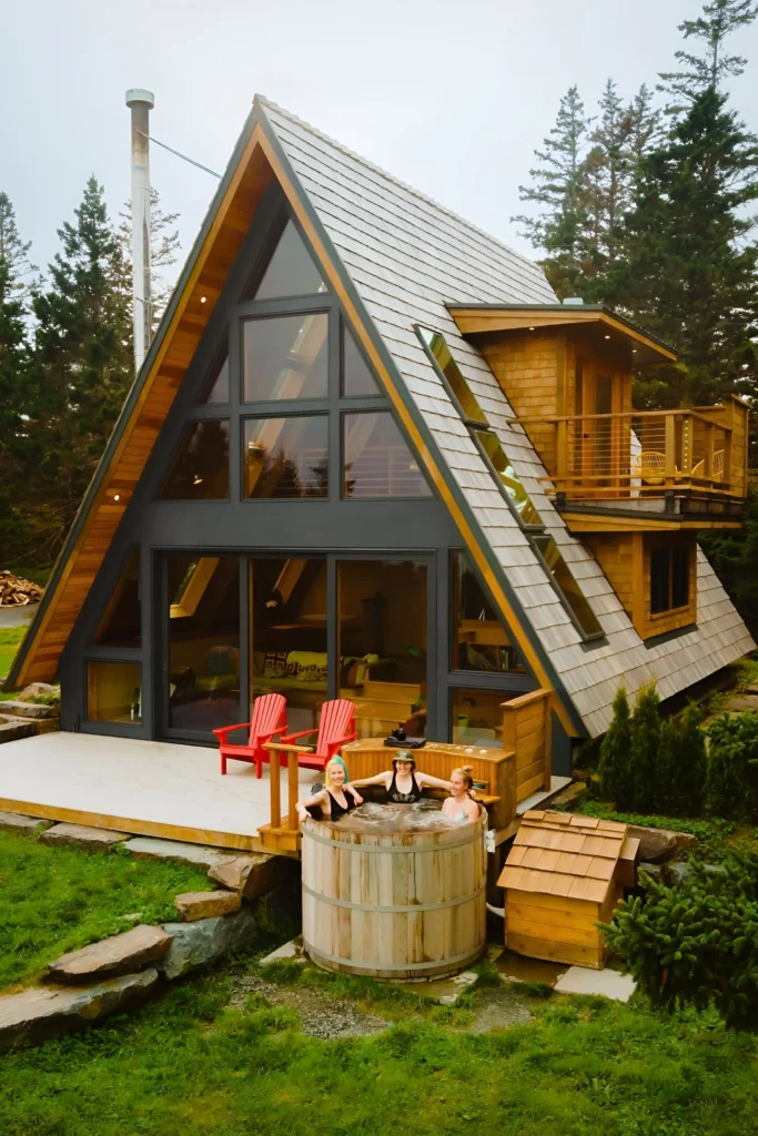 Modern A-frame house with rustic wooden siding, large glass windows, outdoor hot tub, Adirondack chairs, and a second-story balcony in a green setting.