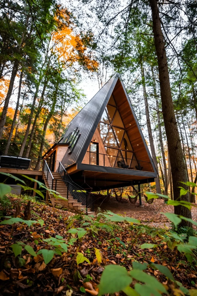 Modern A-frame house with a glass facade, elevated design, wraparound deck, and hammocks, surrounded by tall trees and forest greenery.