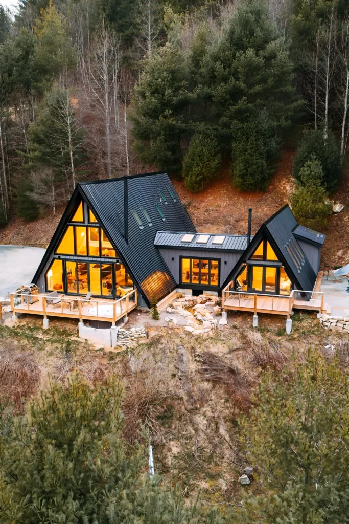 Dual modern A-frame house with black exterior, warm interior lighting, multiple decks, skylights, and a natural hillside setting.