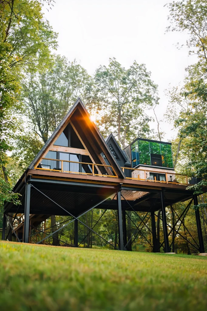 Elevated A-frame house with steel supports, wraparound deck, and a modern glass addition, surrounded by lush forest trees.