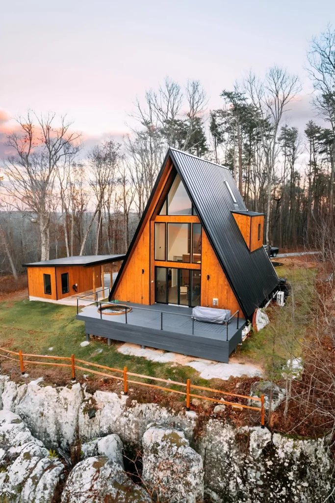 Modern A-frame house with black metal roofing, wood paneling, spacious deck, hot tub, and scenic views of rock formations and forest.