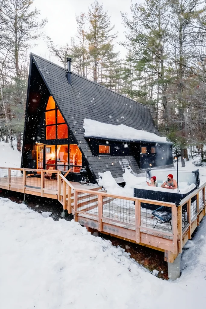 Winter A-frame house with a snow-covered roof, warm glowing interiors, wooden deck, and outdoor hot tub surrounded by a snowy forest.