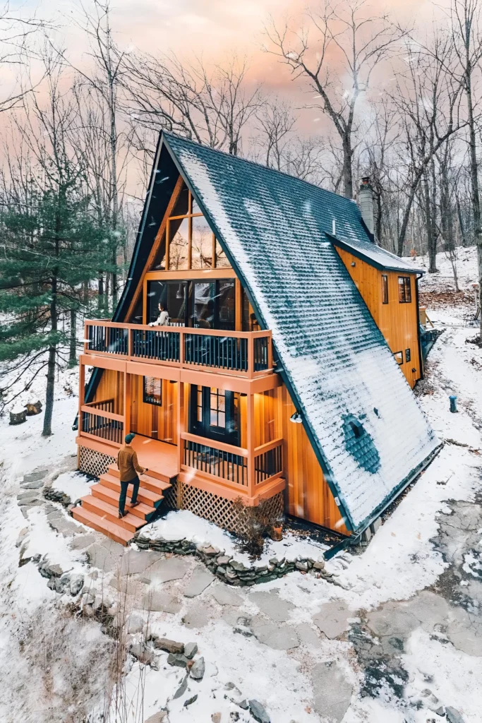 Rustic A-frame house with warm wood siding, two-tiered deck, large windows, and a snow-covered roof in a serene winter setting.