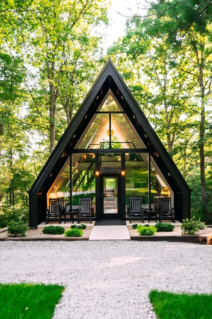 Modern A-frame house with a glass facade, black frame, rocking chairs on the porch, and surrounded by lush green forest.