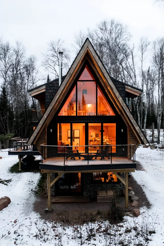 Modern A-frame house with a glass facade, warm glowing interiors, spacious deck, and snowy forest surroundings.