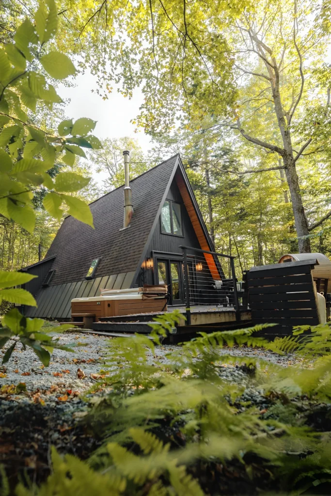 Modern A-frame house with black exterior, outdoor hot tub, wooden deck, and surrounded by vibrant forest greenery.