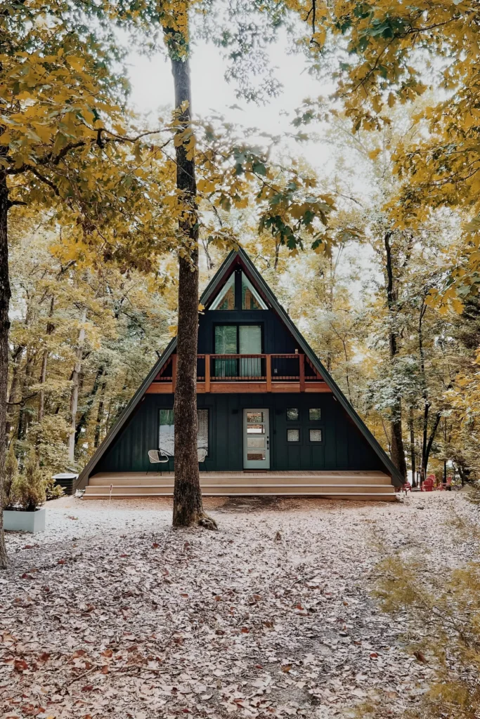 Rustic A-frame house with dark exterior, wooden balcony, and surrounded by vibrant autumn trees in a serene forest setting.