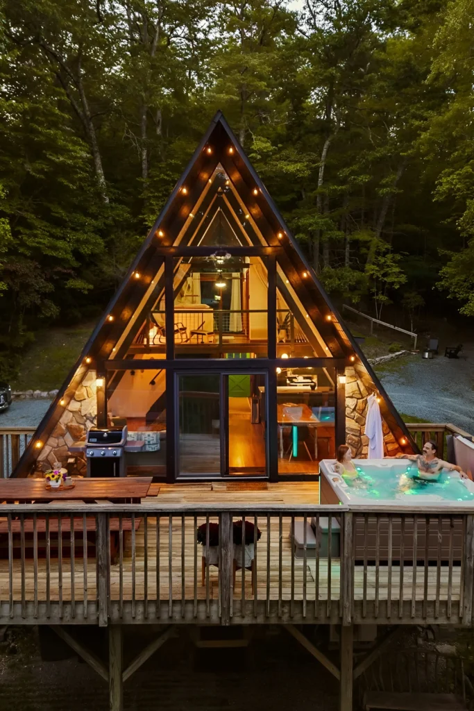 Modern A-frame house with illuminated string lights, spacious deck, hot tub, outdoor dining area, and glass-paneled facade surrounded by lush greenery.