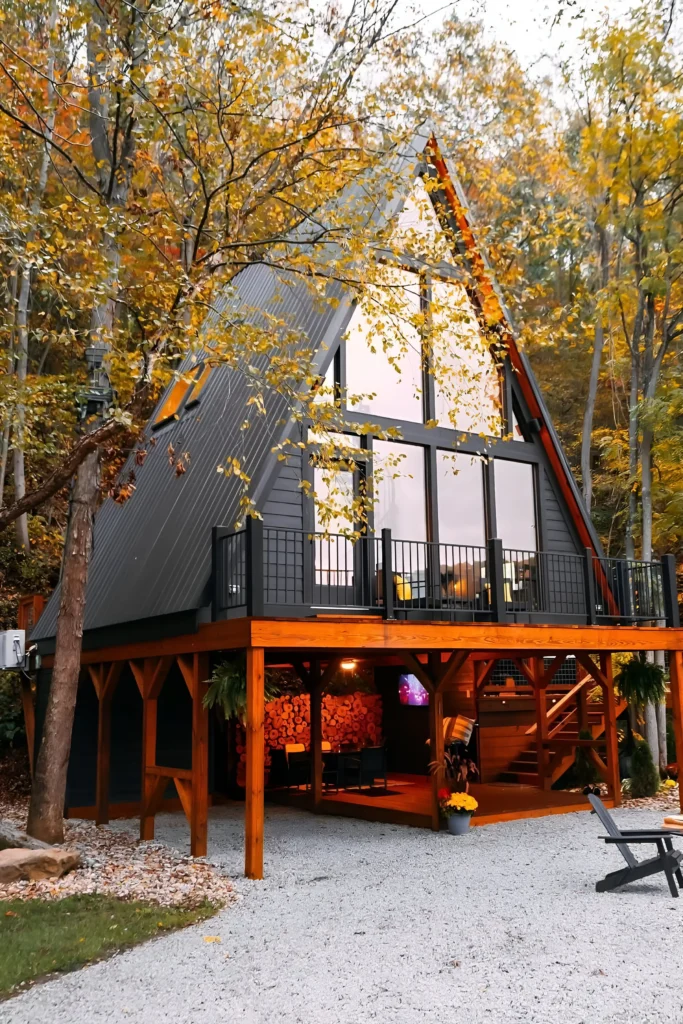 Modern A-frame cabin with black exterior, elevated deck, expansive glass windows, and a cozy lower-level lounge surrounded by autumn trees.