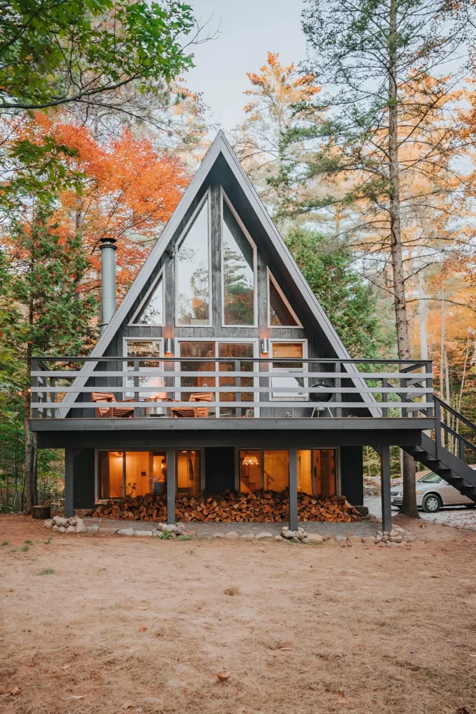 Elevated A-frame cabin with a glass facade, upper-level balcony, and vibrant autumn foliage in a serene forest setting.