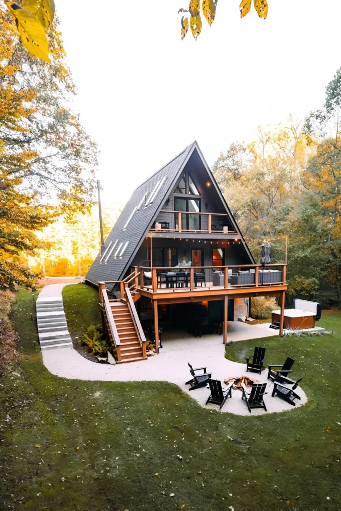 Modern A-frame cabin with upper-level deck, hot tub, fire pit, and lush landscaping surrounded by autumn trees.