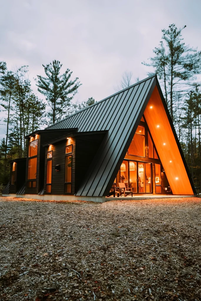 Modern A-frame cabin with sleek black exterior, glowing interior lights, and large glass windows in a serene forest setting.