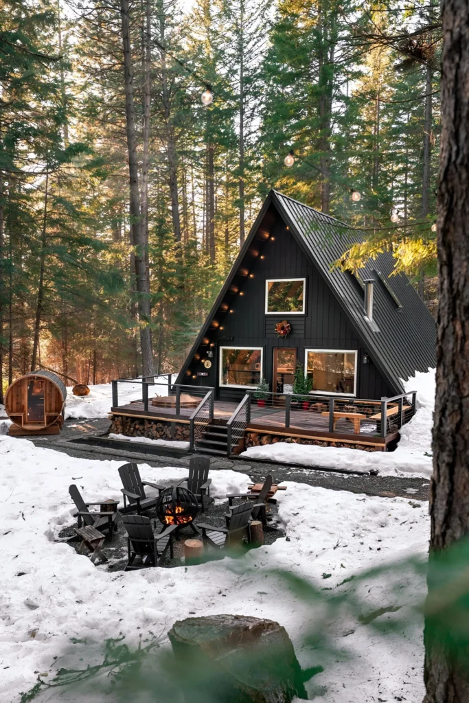 Black A-frame cabin surrounded by snow, with a deck adorned with festive decor, fire pit, and a barrel sauna in a serene forest setting.