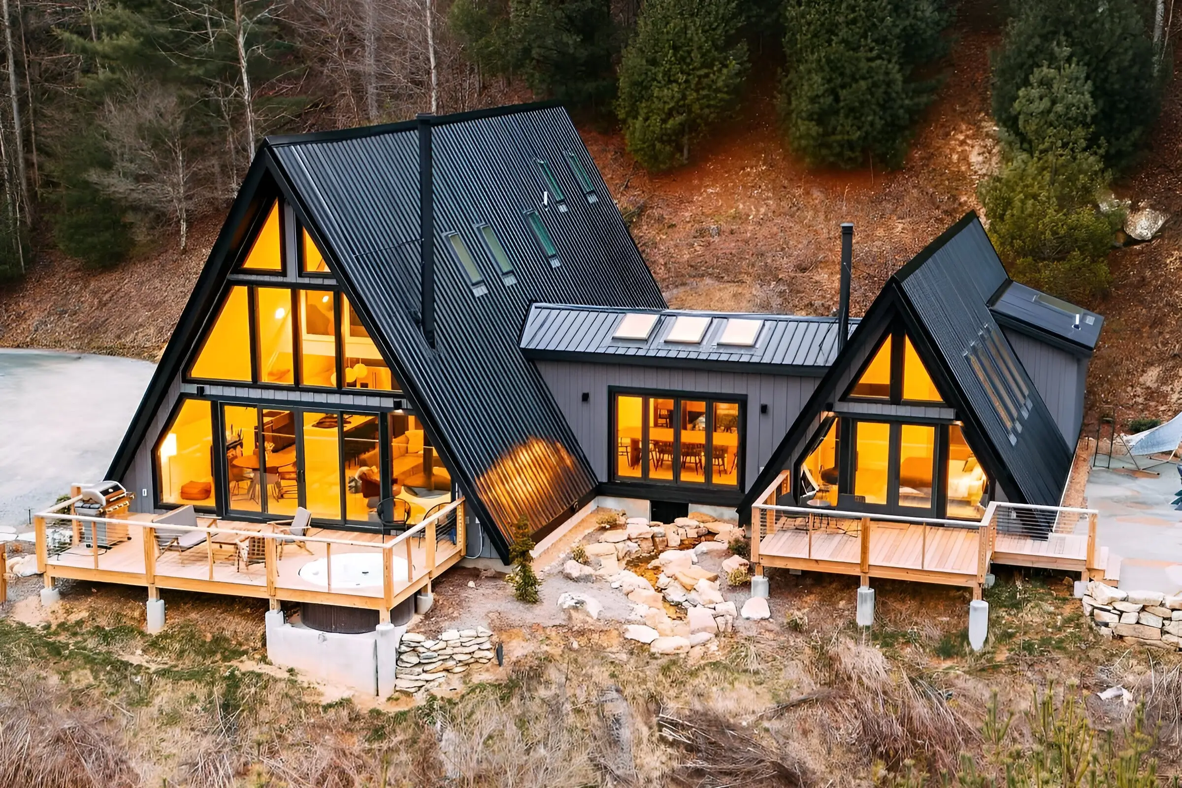 Dual modern A-frame house with black exterior, warm interior lighting, multiple decks, skylights, and a natural hillside setting.