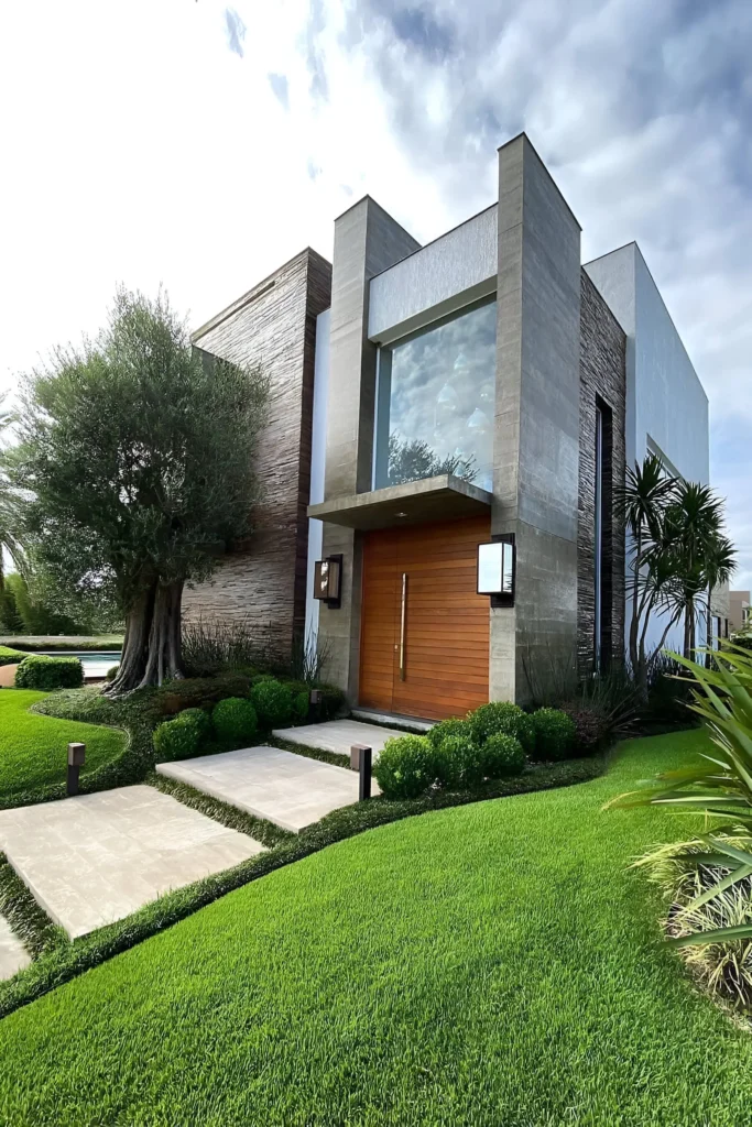 Modern minimalist house featuring textured stone, a wooden entrance door, a large glass panel, and lush landscaping with manicured greenery and an elegant tree.