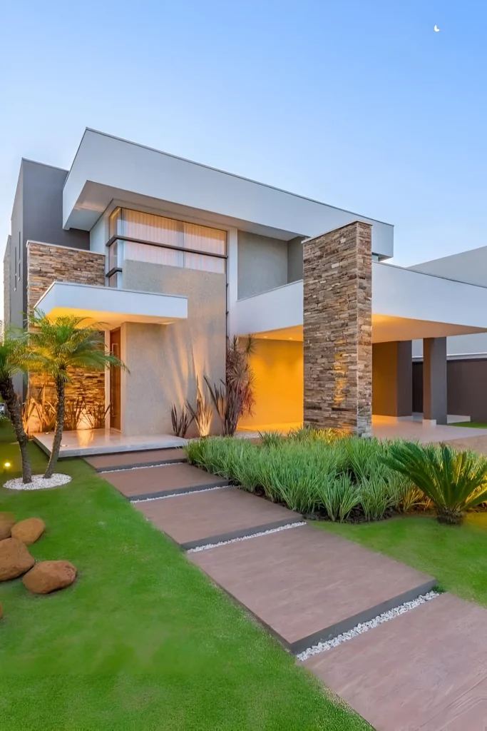 Contemporary home with stone columns, soft lighting, landscaped pathway, and lush greenery for a sophisticated yet natural look.