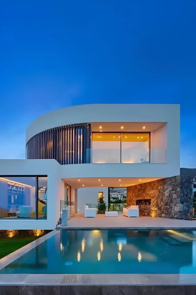 Modern curved villa with a reflective pool, stone-accented walls, and warm evening lighting.