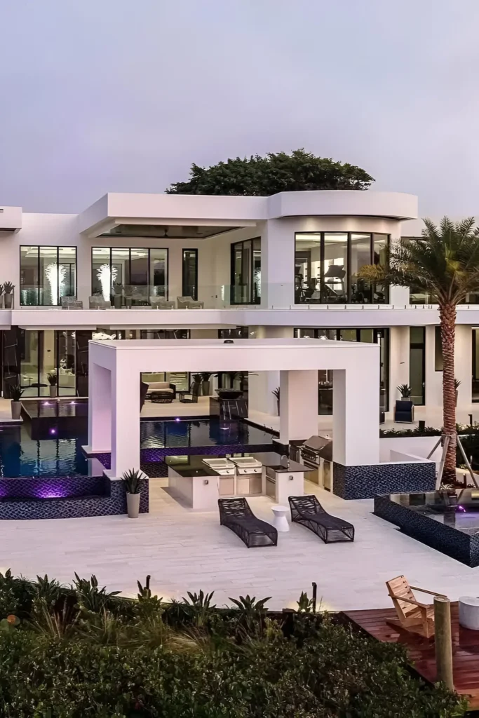 Modern mansion with an expansive pool, outdoor kitchen, vibrant lighting, and sleek glass walls.