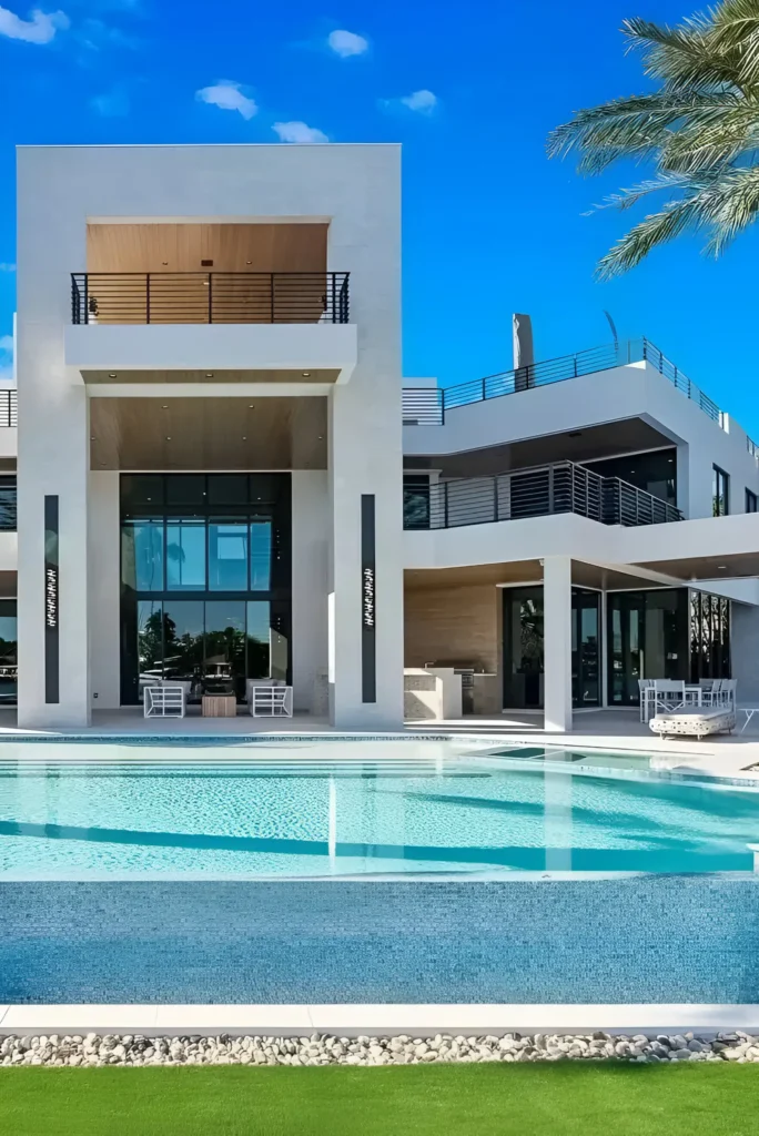 Contemporary luxury mansion with an infinity pool, glass walls, and sleek design, surrounded by palm trees and a vibrant blue sky.